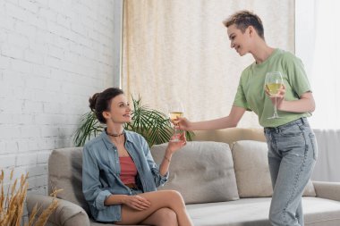 happy pangender person giving glass of white wine to partner sitting on sofa