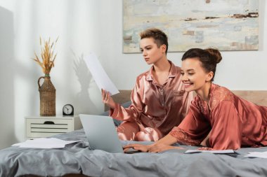 Çift cinsiyetli mutlu kişi yatak odasında kağıtlarla çalışan partnere yakın laptop kullanıyor.