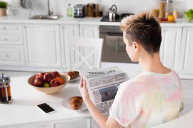 young bigender person reading travel life newspaper near fruits, croissants and smartphone 