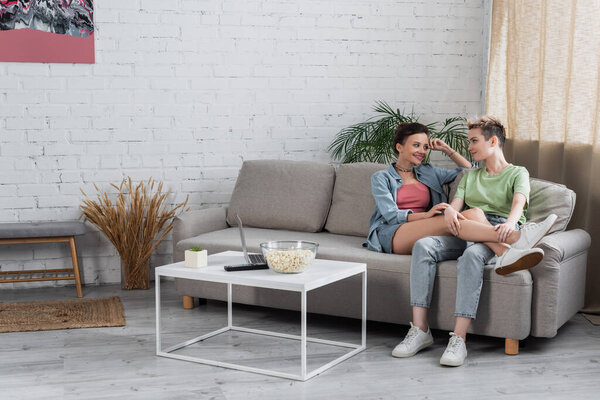 smiling bigender couple talking on couch in living room near laptop and popcorn on coffee table