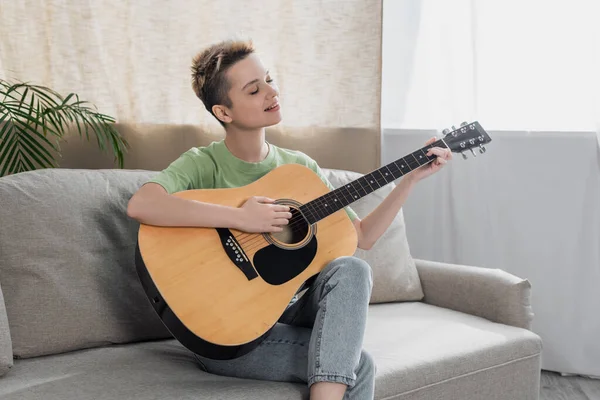 smiling bigender person with short hair sitting on couch and playing acoustic guitar