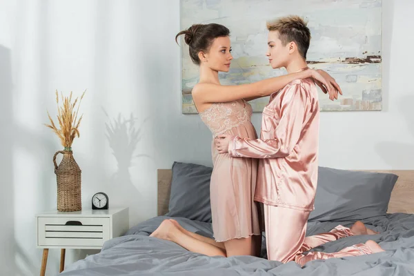 side view of pangender people in silk pajamas and nightdress looking at each other on bed
