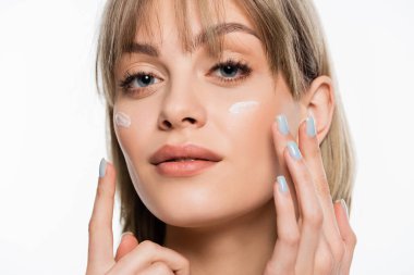 Kâküllü kadın yüzüne krem sürüyor ve beyaz kameraya bakıyor.