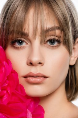 close up of young woman with bangs near bright pink decorative flower isolated on white