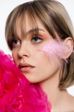 pretty young woman with feather on cheek near bright pink decorative flower isolated on white