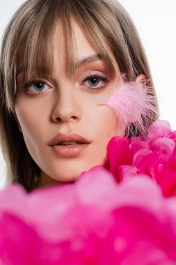 pretty young woman with feather on cheek near blurred pink decorative flowers isolated on white
