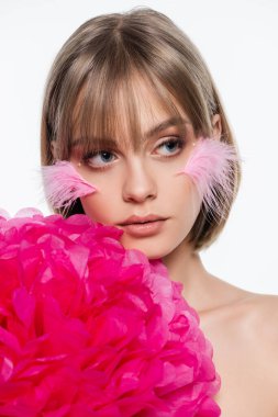 young woman with decorative elements in makeup and pink feathers on cheeks near bright flower isolated on white