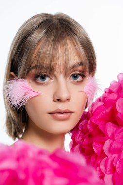 young woman with decorative elements in makeup, bangs and pink feathers on cheeks near flowers isolated on white