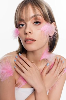 young woman with decorative elements and pink feathers on face and body posing with crossed arms isolated on white