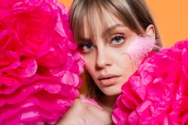 young woman with decorative beads in makeup and feathers on cheeks near pink flowers isolated on orange