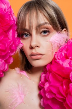 woman with decorative beads in makeup and pink feathers on cheeks near flowers isolated on orange