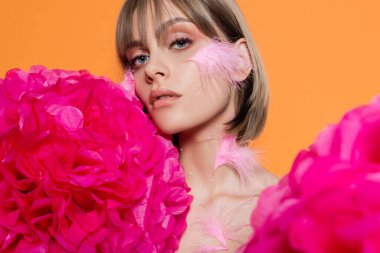 young woman with decorative beads in makeup and pink feathers on cheeks near flowers isolated on orange