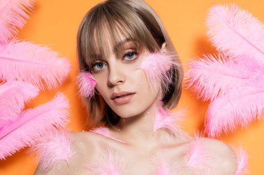 young woman with decorative beads in makeup near pink feathers isolated on orange