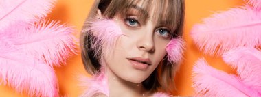 young woman with decorative beads in makeup near pink feathers isolated on orange, banner
