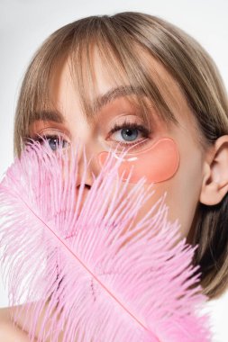 young woman with hydrogel eye patches covering face with soft pink feather isolated on white