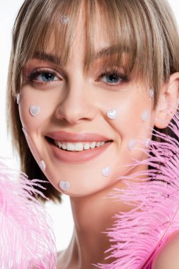 close up of happy woman with nacreous heart shape elements on cheeks near pink feathers isolated on white 
