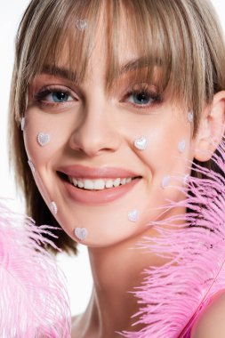 close up of happy woman with nacreous heart shape elements on cheeks near pink feathers isolated on white  clipart