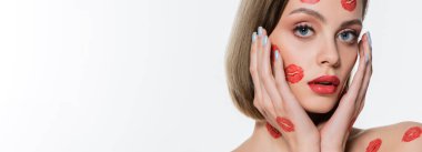 young woman with red kiss prints on cheeks touching face isolated on white, banner