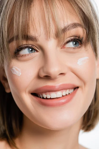 close up of happy woman with bangs and cream on cheeks isolated on white