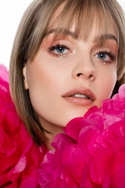 close up of young woman with bangs and rhinestones under blue eyes near pink flowers isolated on white
