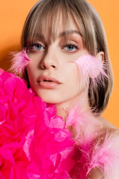 young woman with decorative beads in makeup and feathers on cheeks near bright pink flower isolated on orange