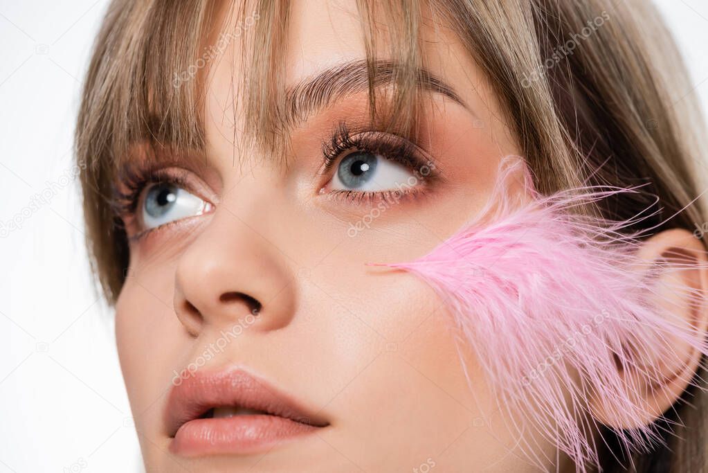 close up of sensual young woman with blue eyes and pink feather on face looking up isolated on white