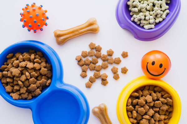 top view of bone shaped pet treats near bowls with dry pet food and rubber toys isolated on white
