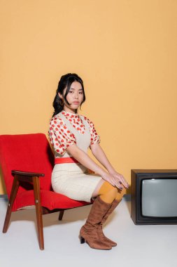 Güzel Asyalı kadın, turuncu arka planda, televizyonun yanındaki retro koltukta oturuyor.
