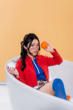 Young asian model in vintage clothes and with glitter on face holding balls while sitting in bathtub on orange background clipart