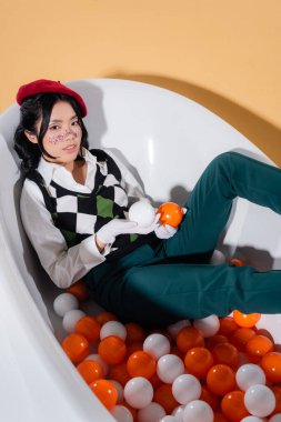 High angle view of stylish asian woman holding balls while sitting in bathtub on orange background clipart