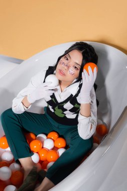 High angle view of trendy asian model in gloves holding balls while sitting in bathtub on orange background clipart