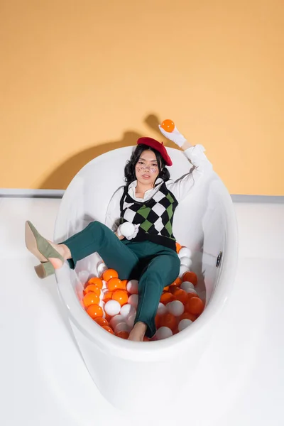 Trendy Asian Model Beret Holding Balls Looking Camera Bathtub Orange — Stock Photo, Image