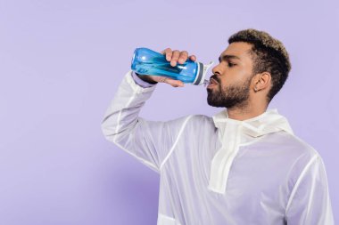 Sakallı Afro-Amerikan sporcusu elinde spor şişesiyle mor üzerine serinletici su içiyor. 