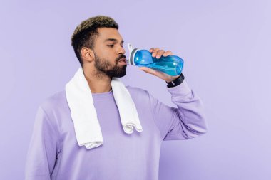Beyaz havluyla dikilip mor üzerine izole edilmiş su içen kazaklı genç Afrikalı Amerikalı adam. 