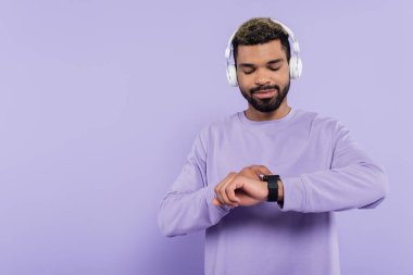 Kablosuz kulaklıklı sakallı Afro-Amerikan adam mor üzerine izole edilmiş akıllı saate bakıyor.