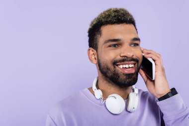 Kablosuz kulaklıklı mutlu Afro-Amerikan adam mor renkte izole edilmiş akıllı telefonuyla konuşuyor. 