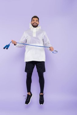 Mutlu Afro-Amerikan bir adam spor giyim kuşamında havada asılı duruyor ve elinde mor iplerle atlıyor.