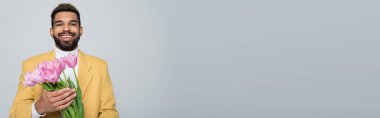 Mutlu Afro-Amerikan erkeği şık giyinmiş elinde gri pankartta izole edilmiş pembe laleler tutuyor.