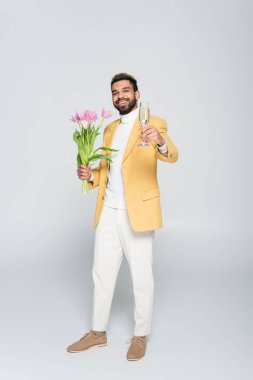 Sarı ceketli, elinde pembe laleler ve gri üzerinde izole edilmiş bir bardak şampanya tutan mutlu Afro-Amerikan bir adam. 
