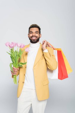 Sarı ceketli, polo yakalı, pembe laleli ve gri renkli alışveriş torbalı Afro-Amerikan bir adam. 