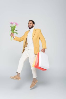 Sarı ceketli, polo yakalı, pembe laleli ve gri çantalı neşeli Afro-Amerikan bir adam. 