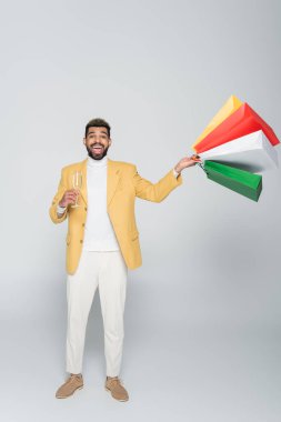Sarı ceketli neşeli Afro-Amerikalı adam elinde poşetler ve gri üzerine bir bardak şampanya tutuyor.