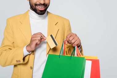 Sarı ceketli, elinde alışveriş çantası ve kredi kartı olan neşeli Afro-Amerikan adamın gri üzerinde izole edilmiş görüntüsü. 