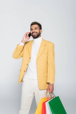 Sarı ceketli ve polo yakalı mutlu Afro-Amerikan bir adam alışveriş torbaları tutuyor ve gri renkli telefondan konuşuyor. 