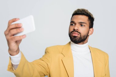 Sarı ceketli neşeli Afro-Amerikalı adam gri renkli telefondan selfie çekiyor. 