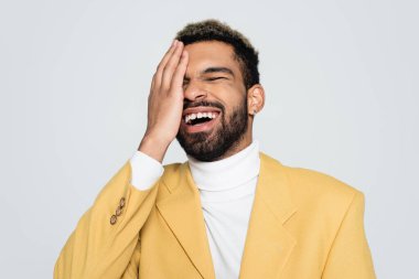 Pastel sarısı ceketli mutlu Afro-Amerikan adam gözlerini kapatıyor ve grinin üstünde gülüyor. 