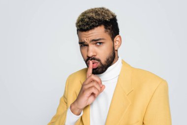 young african american man with blue eyes in stylish yellow blazer showing hush sign isolated on grey 