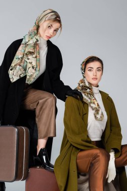 Paltolu ve desenli başörtülü kadınlar gri renkli bavulların yakınındaki kameraya bakıyorlar.