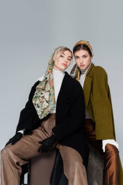 Şık ceketli ve zarif mendilli kadınlar, gri renkte izole bir şekilde otururken kameraya bakıyor.