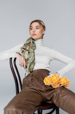 Eşarplı moda kadın sandalyede oturuyor, portakallar ağda, gri çantada.